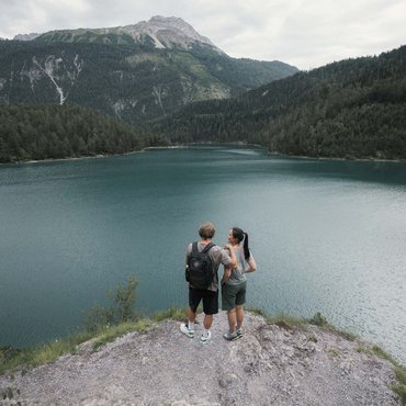 Zwei Wanderer stehen am Seeufer mit Blick auf Berg und Wald