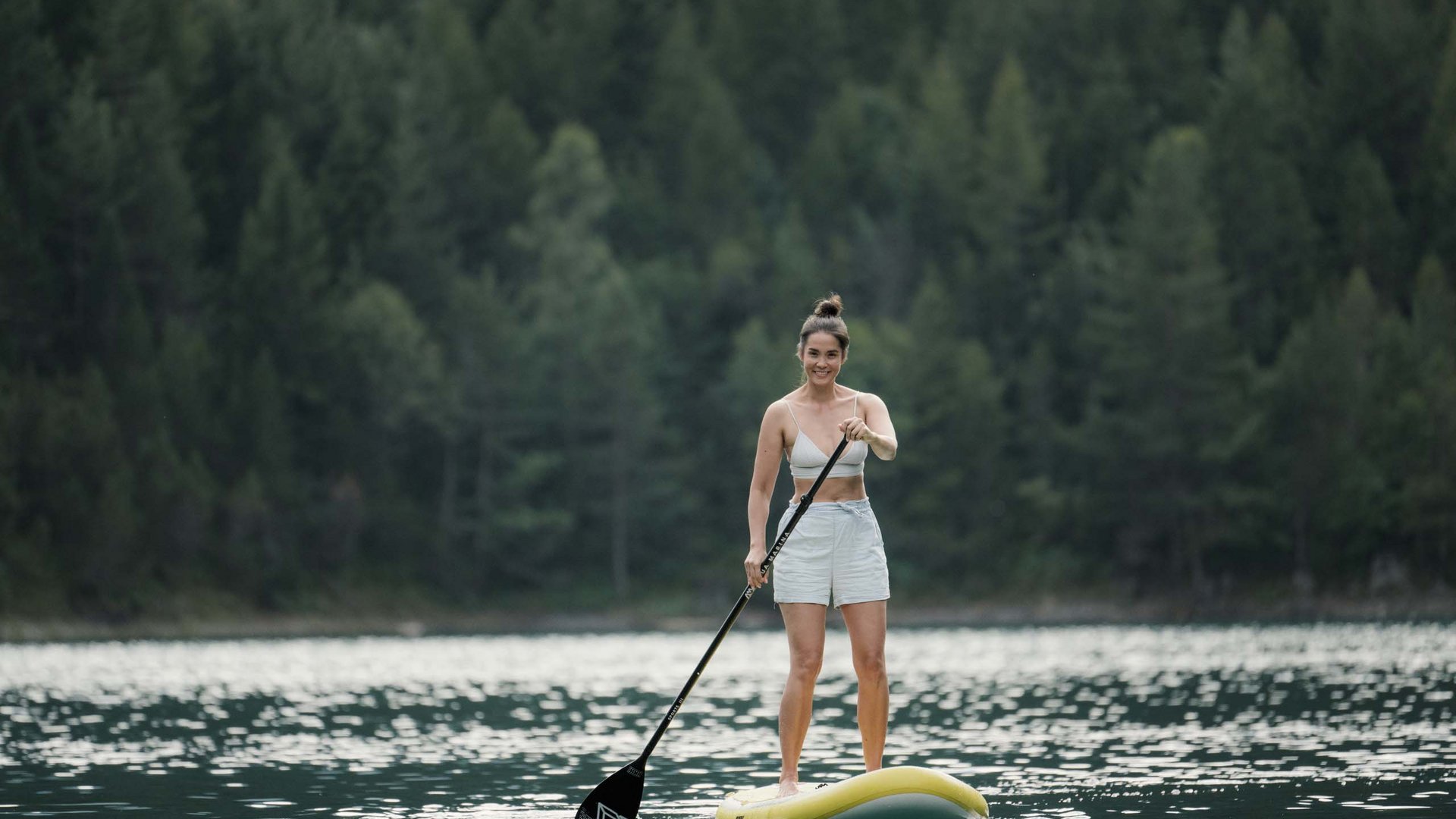 Frau steht auf Stand-Up-Paddleboard auf See vor Waldkulisse