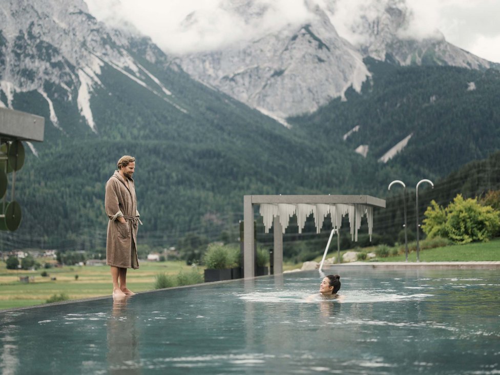 Mann im Bademantel steht am Pool, Frau schwimmt, Berge im Hintergrund