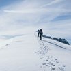 Zwei Wanderer steigen mit Stöcken auf eine schneebedeckte Bergspitze bei blauem Himmel