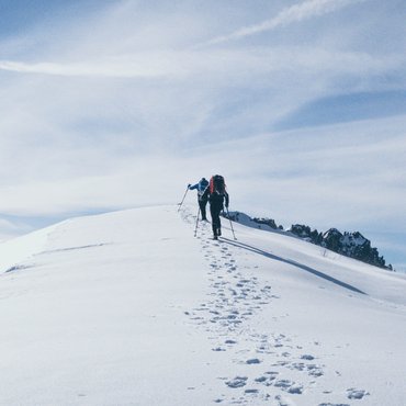 Zwei Wanderer steigen mit Stöcken auf eine schneebedeckte Bergspitze bei blauem Himmel