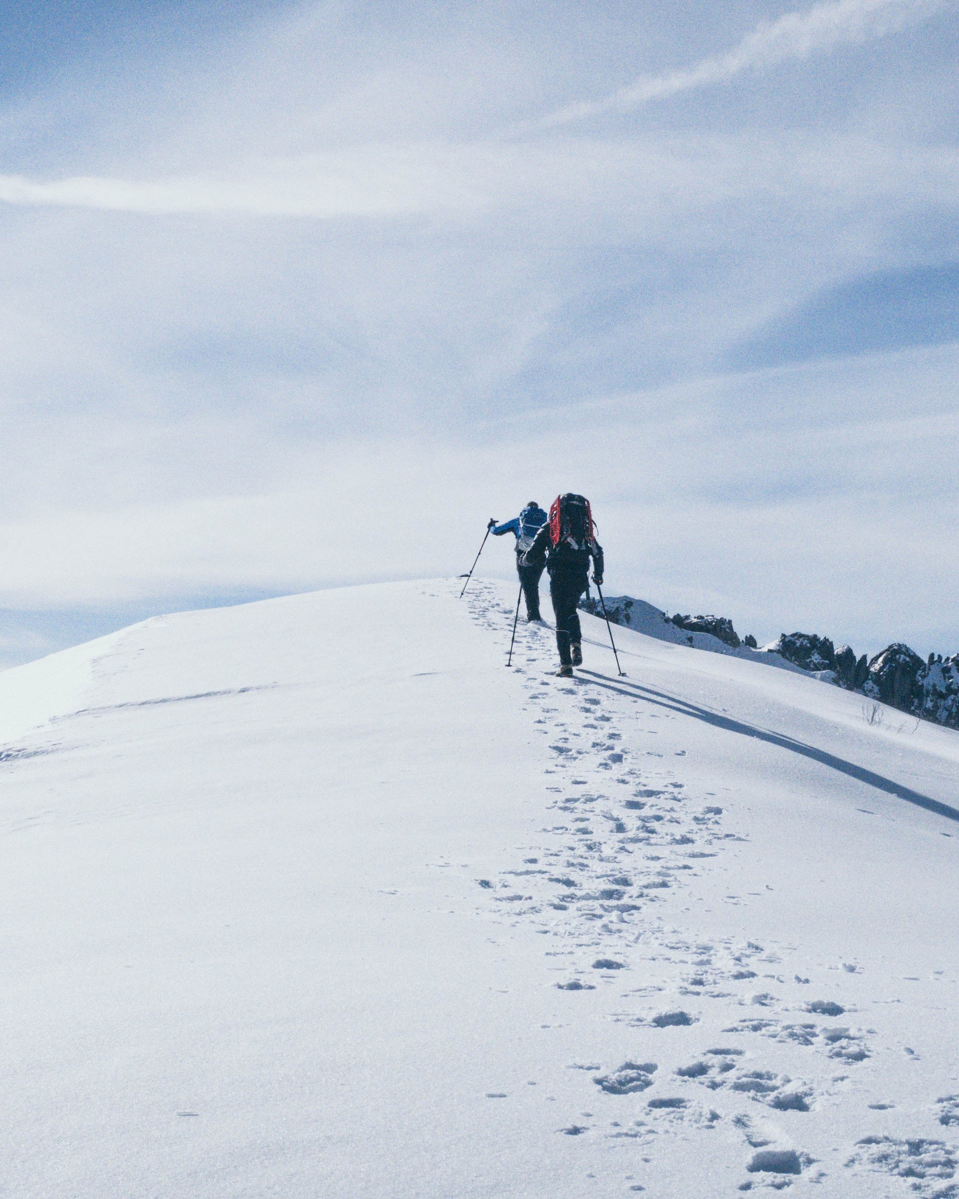 Zwei Wanderer steigen mit Stöcken auf eine schneebedeckte Bergspitze bei blauem Himmel