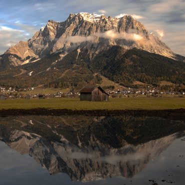 Berg mit Wolken und Spiegelung im Wasser vor einer Hütte und Dorf