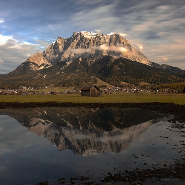 Berg mit Wolken und Spiegelung im Wasser vor einer Hütte und Dorf