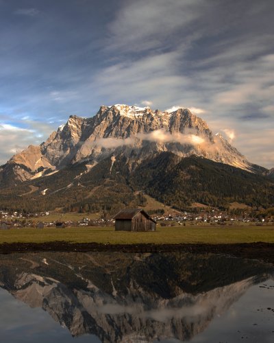 Berg mit Wolken und Spiegelung im Wasser vor einer Hütte und Dorf