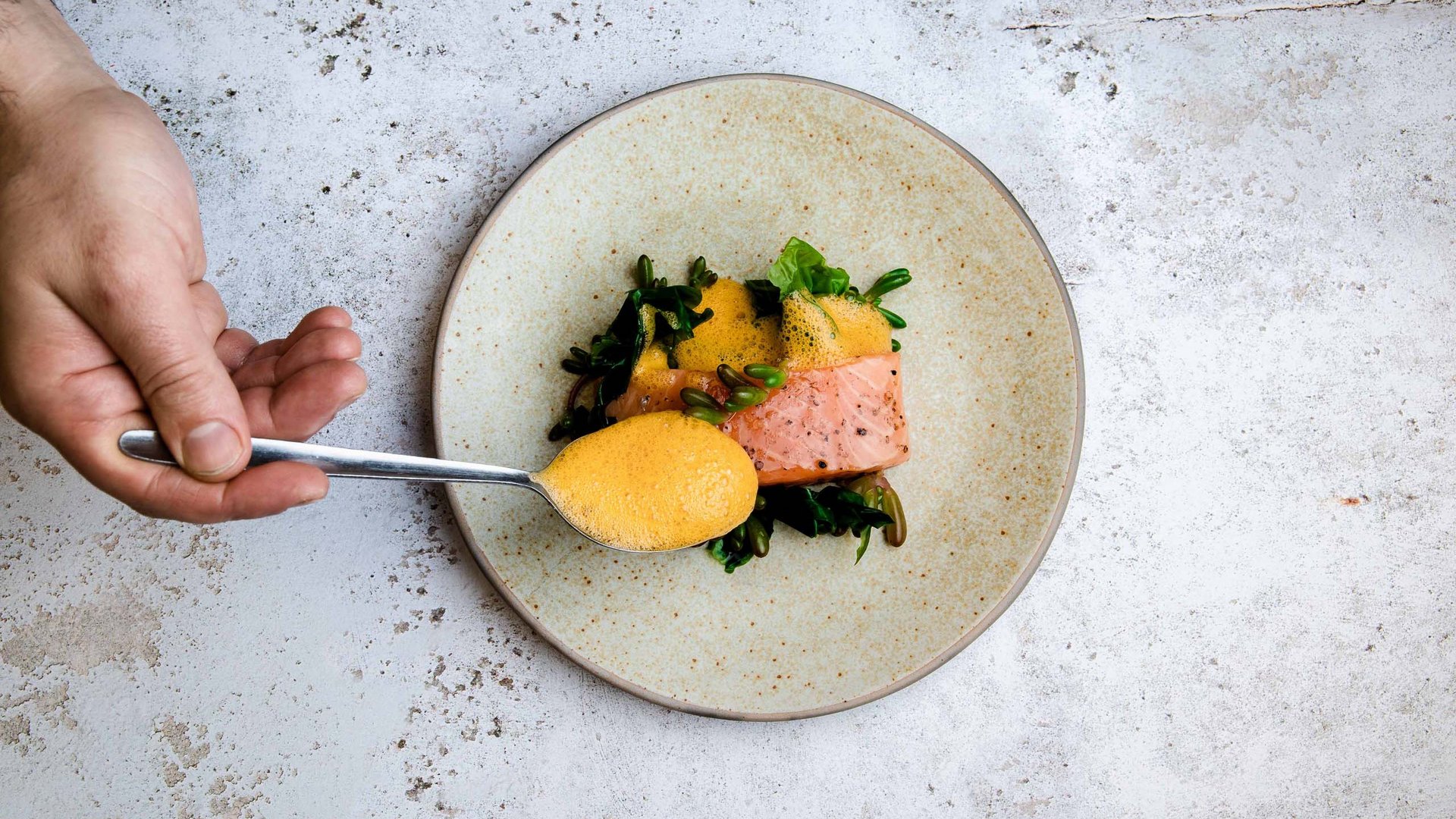 Hand gibt Löffel mit Sauce über Lachs und Gemüse auf Teller