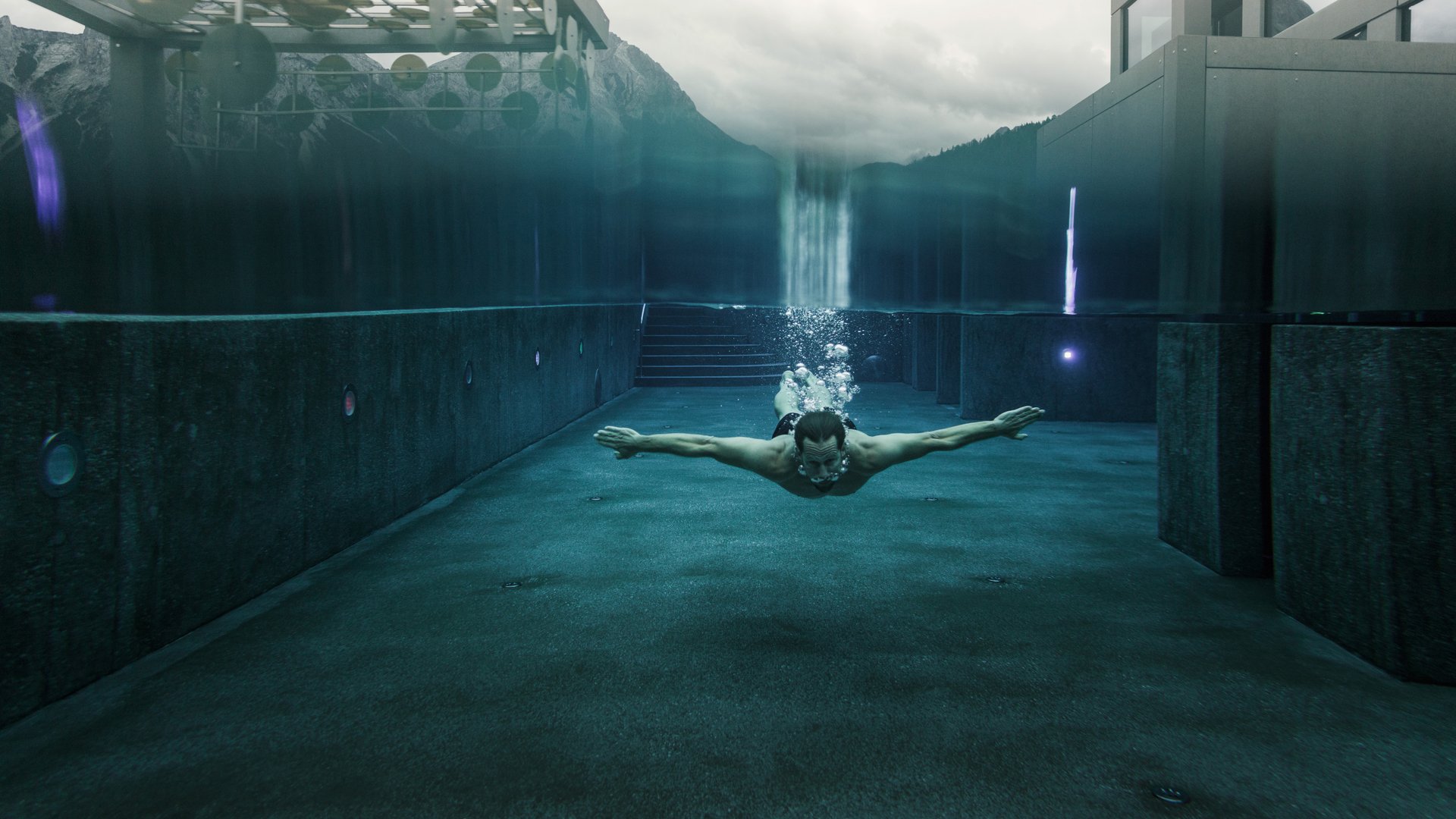 Mann schwimmt unter Wasser in modernem Schwimmbecken mit Bergblick
