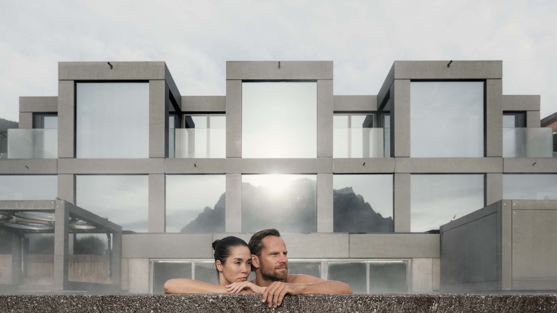 Paar entspannt in beheiztem Außenpool vor modernem Gebäude mit Bergblick