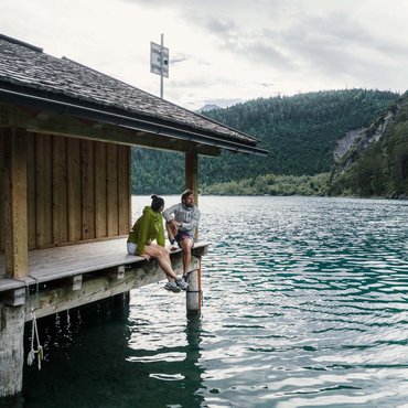 Zwei Personen sitzen auf einem Steg an einem See mit Berglandschaft im Hintergrund.