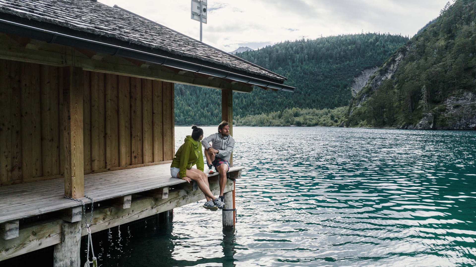 Zwei Personen sitzen auf einem Steg an einem See mit Berglandschaft im Hintergrund.