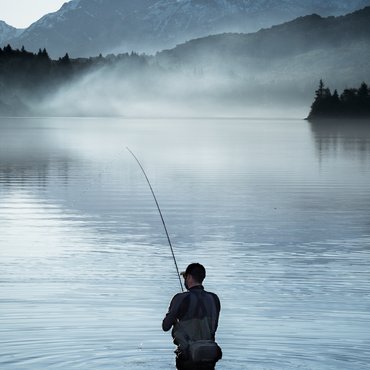 Angler steht im ruhigen See vor schneebedeckten Bergen bei Morgennebel