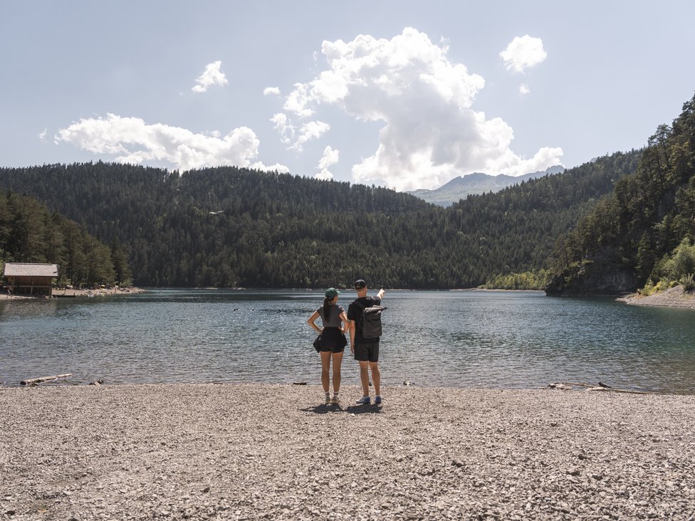 Zwei Personen stehen am Kiesstrand und schauen auf einen See mit bewaldeten Bergen