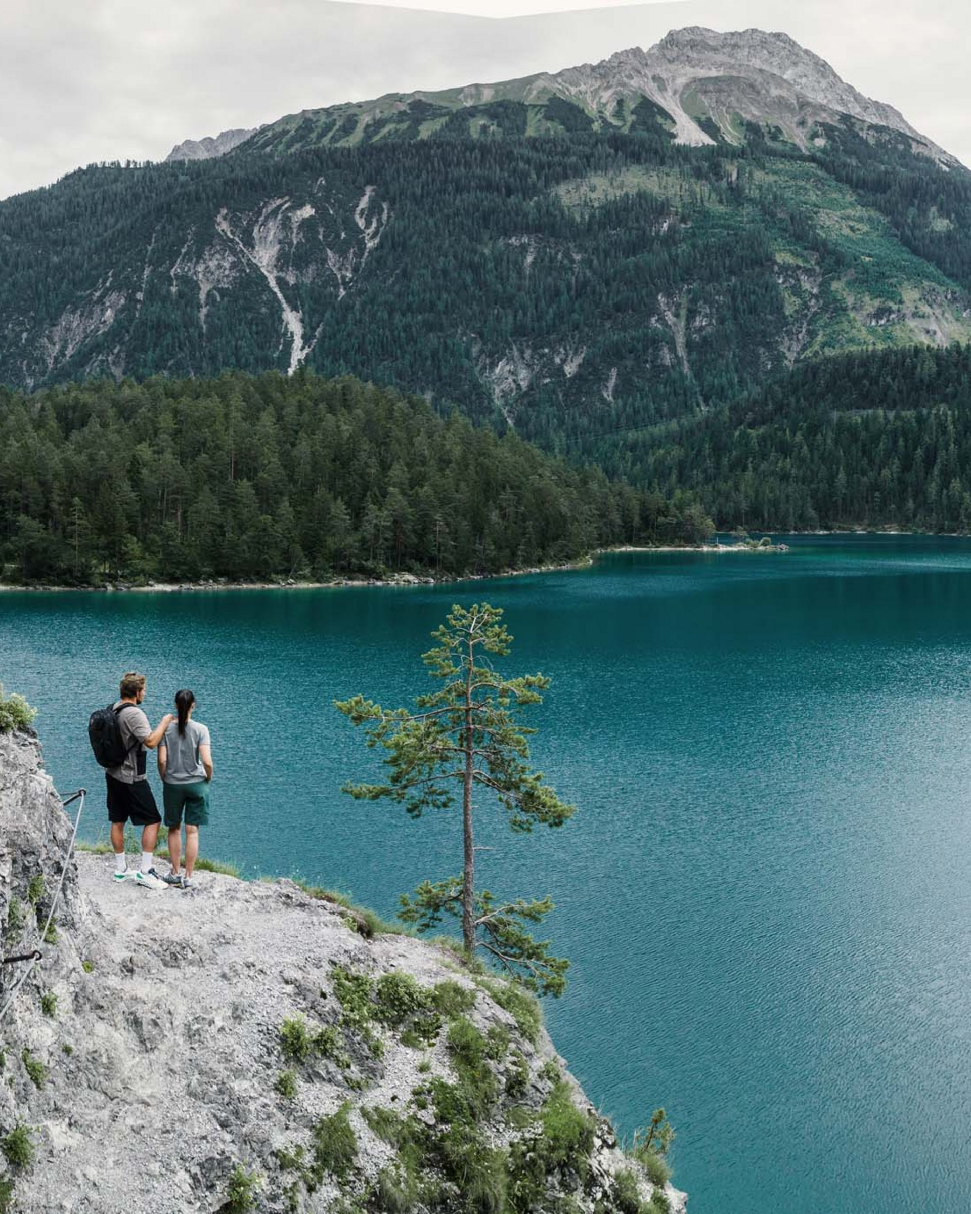 Zwei Personen wandern an einem See mit Bergen und Wald im Hintergrund