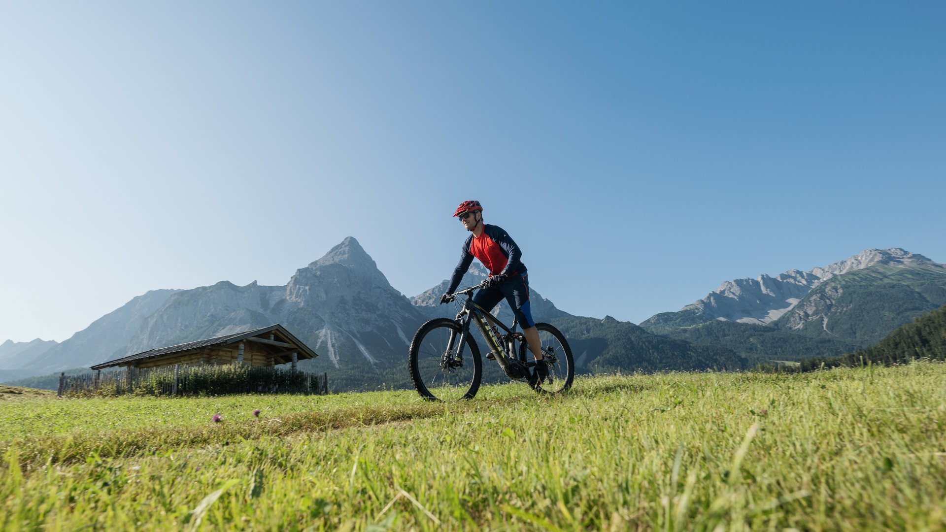 Mann fährt Mountainbike auf einer Wiese mit Bergkulisse bei klarem Himmel