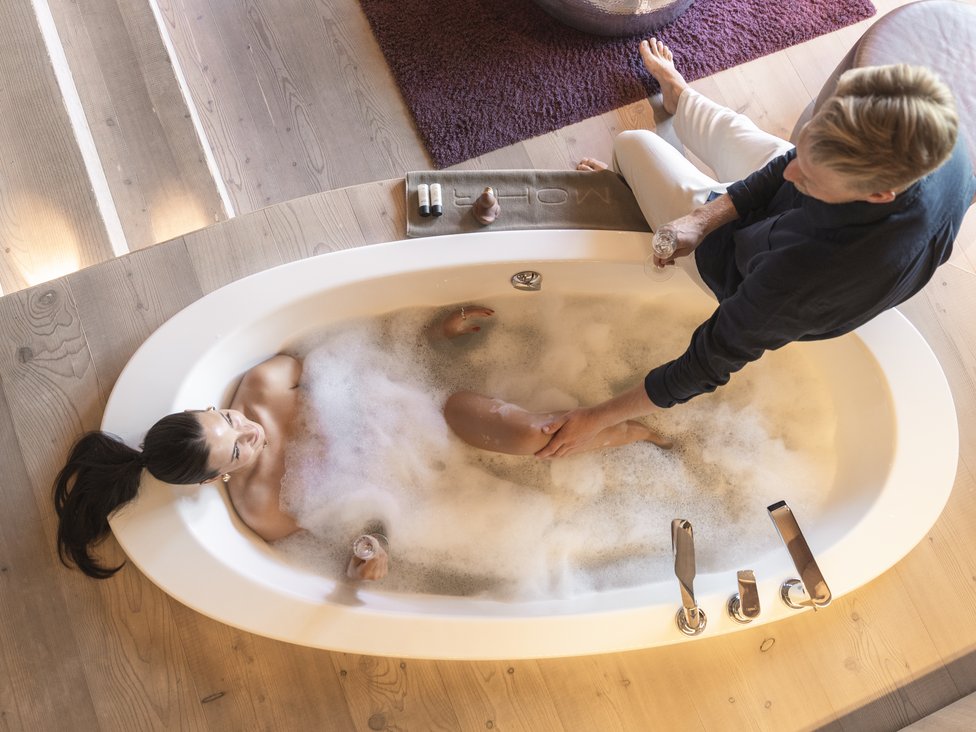 Frau entspannt in Schaumbad, Mann sitzt neben Badewanne und hält ihr Bein