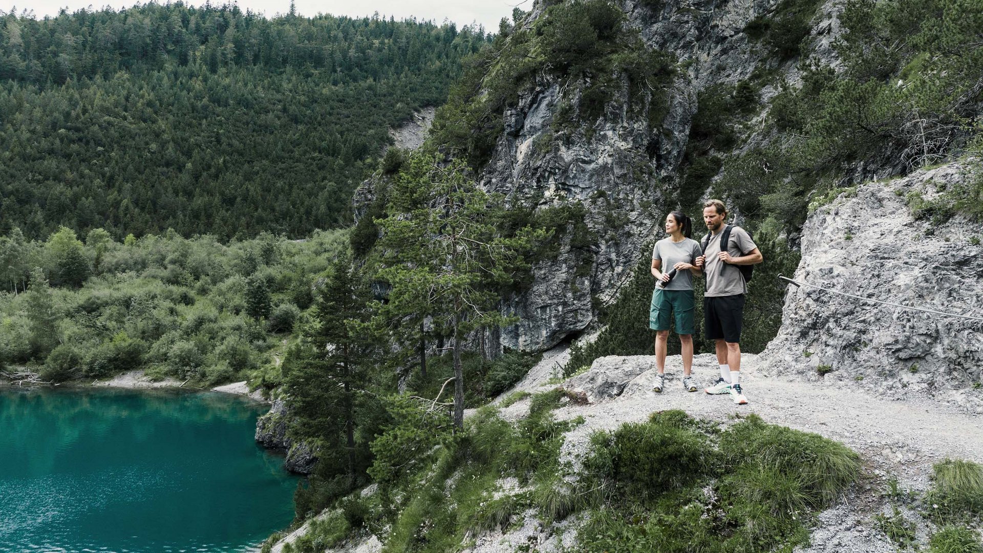 Zwei Wanderer stehen auf einem Felsen mit Blick auf einen Bergsee und Wald