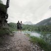 Zwei Menschen wandern an einem Bergsee mit bewaldeten Bergen im Hintergrund
