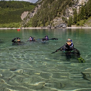 Vier Taucher im klaren Bergsee vor bewaldetem Felsen