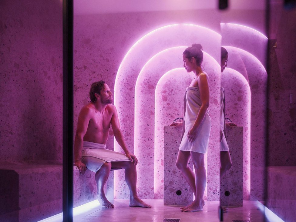 Großzügig und exklusiv: Ihr Wellnessurlaub in Tirol Mann sitzt, Frau steht in Spa mit rosa Neonlicht und Handtüchern