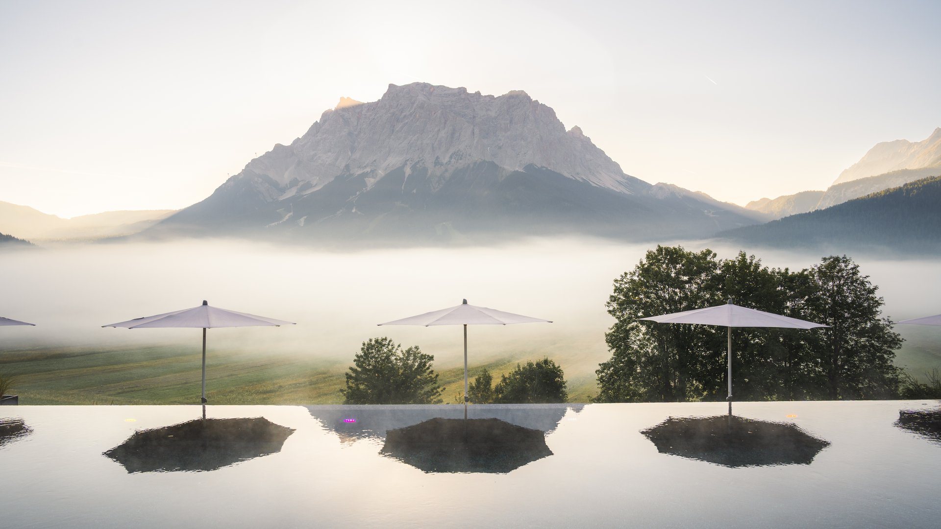 Großzügig und exklusiv: Ihr Wellnessurlaub in Tirol Infinity-Pool mit Sonnenschirmen und Bergpanorama im Morgennebel