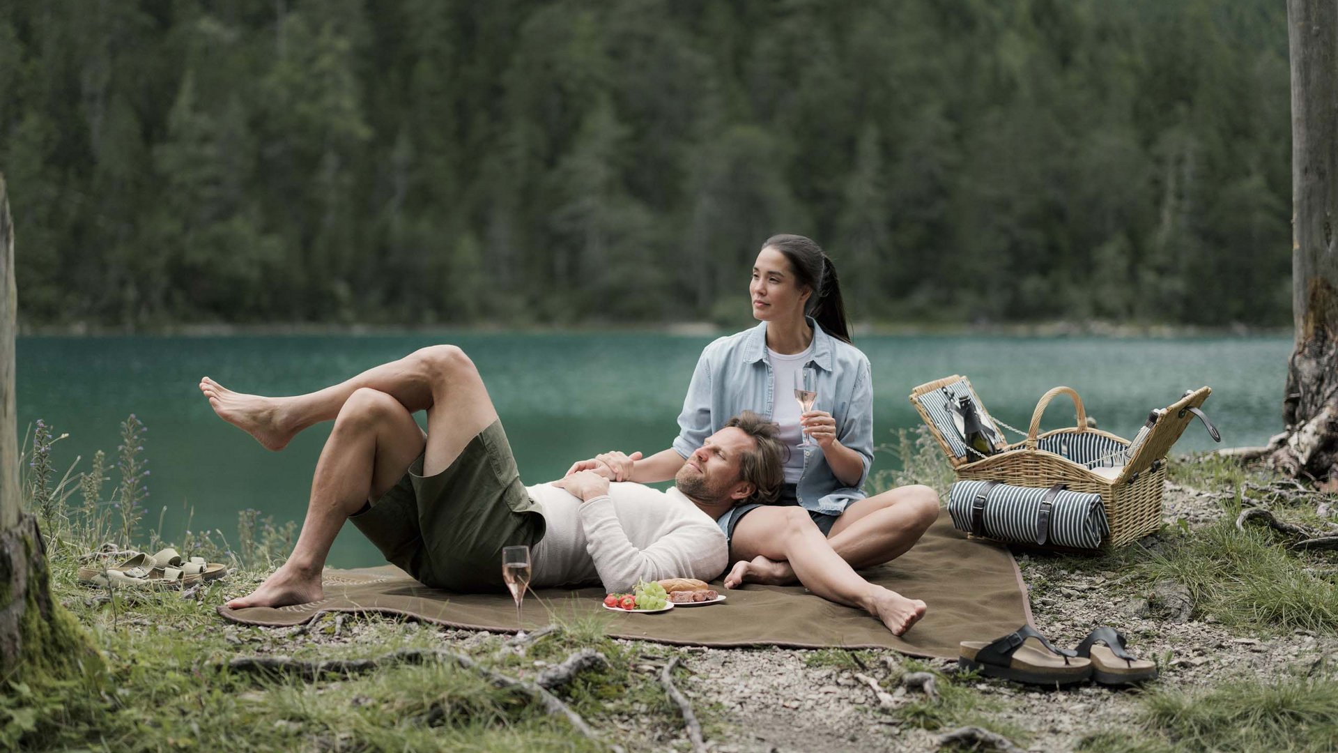 Paar macht Picknick am See mit Wein und Snacks auf Decke im Wald