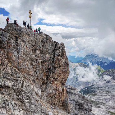 Berggipfel mit Wanderern und Gipfelkreuz unter bewölktem Himmel