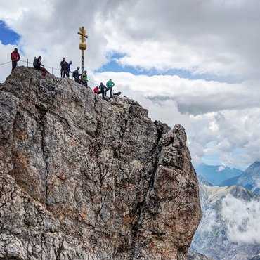 Berggipfel mit Wanderern und Gipfelkreuz unter bewölktem Himmel