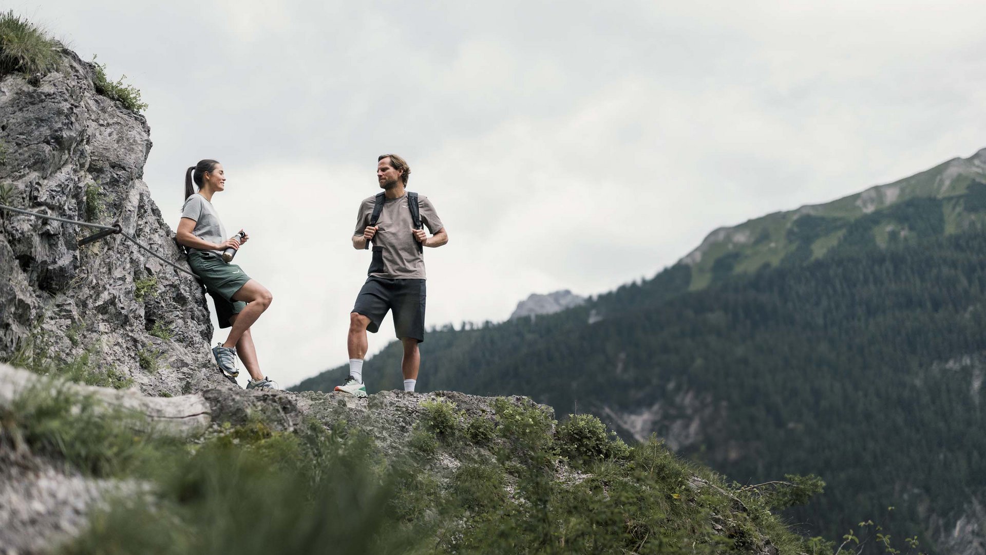 Zwei Wanderer machen Pause auf felsigem Bergweg mit Wald im Hintergrund