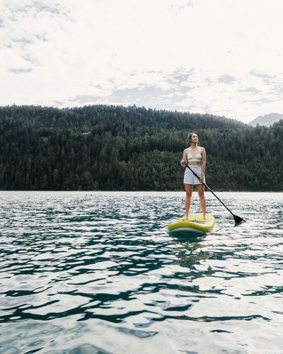 Frau steht auf Paddleboard auf See vor bewaldeten Bergen
