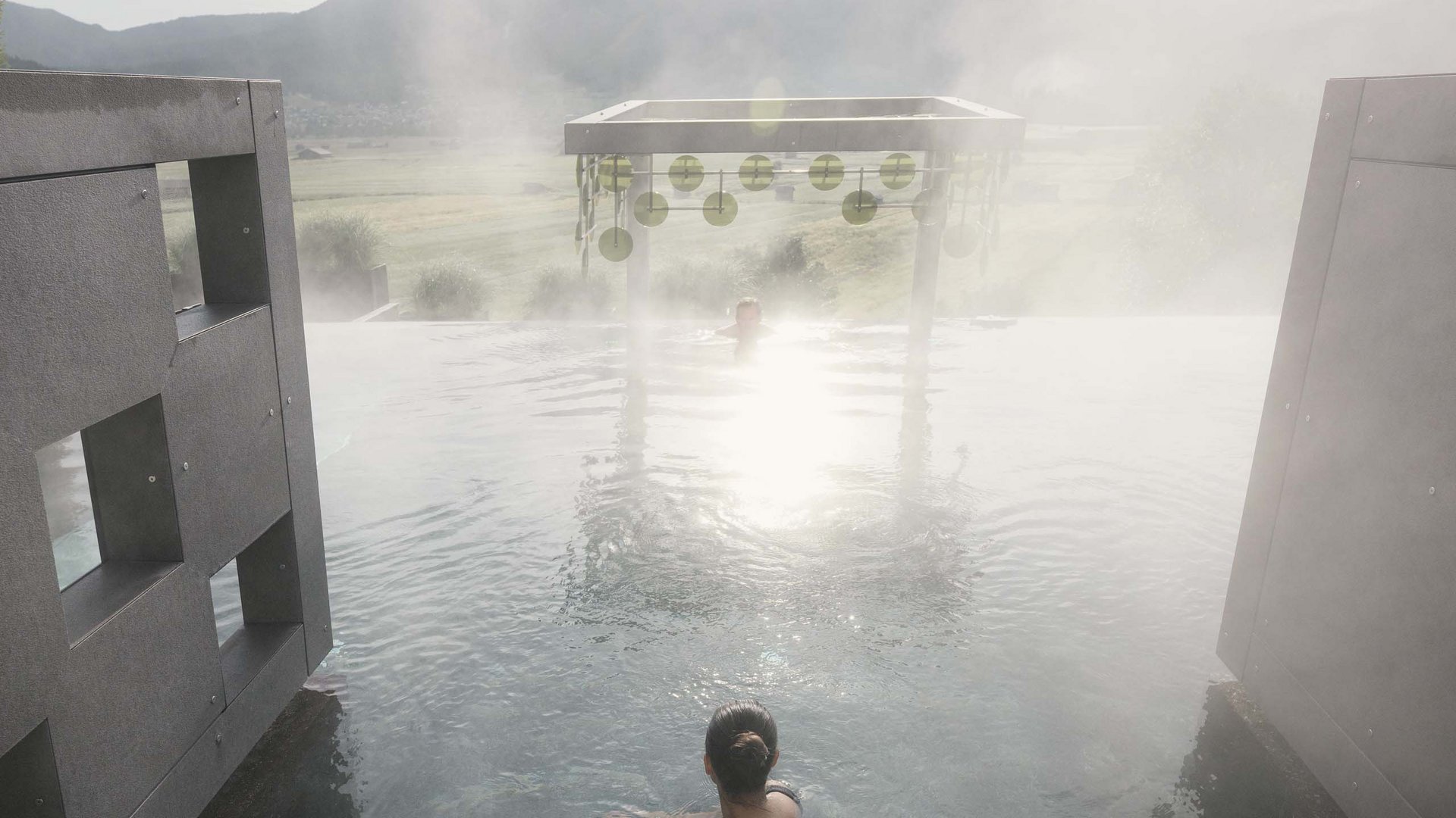 Person betritt dampfendes Außenbecken mit Bergpanorama im Sonnenlicht